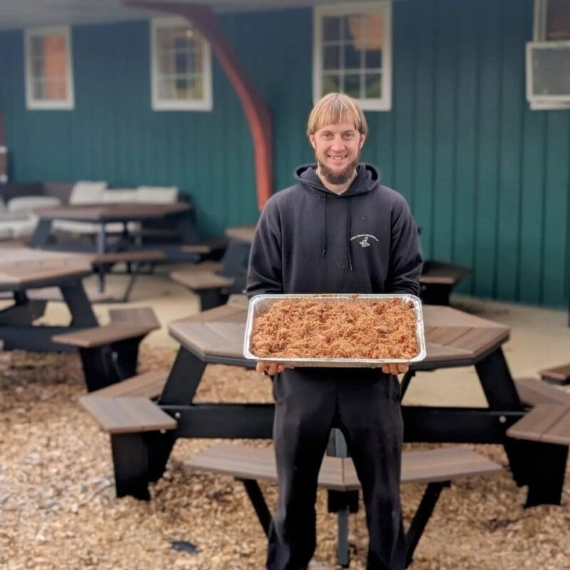Smiling man in a black hoodie holding a large aluminum tray of pulled pork outside a BBQ restaurant patio with picnic tables.